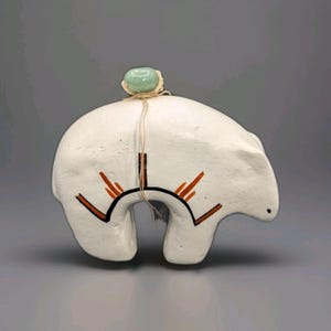 May include: A white ceramic bear figurine featuring a smooth matte finish. The bear is decorated with black and orange painted geometric lines. A light green polished stone is tied to the bear's back with thin, light-colored twine.