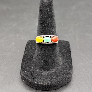 May include: A silver ring with three oval-shaped stones in yellow, green, and red. The ring is displayed on a black velvet ring holder against a dark background. The stones are set in a row.