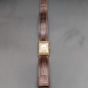 May include: A rectangular gold-tone wristwatch featuring a textured, off-white dial with black numerals and a small sub-dial. The watch is attached to a dark brown, embossed leather strap with a gold-tone metal buckle and a matching metal keeper.