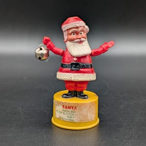 May include: A vintage Santa Claus push-button toy standing on a yellow cylindrical base. The figure wears a red suit with white trim, a black belt, and black boots. Santa holds a small silver jingle bell in one hand. A label on the base reads SANTA PUSH BUTTON Made by Kohner Bros.