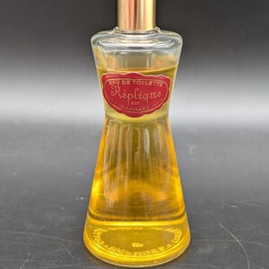 May include: A vintage perfume bottle with a gold cap and a clear glass body. The bottle contains a golden-yellow liquid and features a red label with the text "Eau de Toilette Réplique". The bottle has a unique hourglass shape.