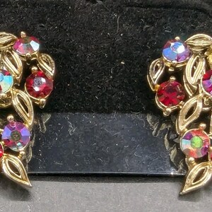 May include: A pair of vintage gold-toned earrings. Each earring features a leaf design with red and iridescent crystal accents. The earrings are set against a black background.