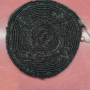 May include: A circular coin purse covered in small, shiny black beads arranged in a concentric circular pattern. The purse features a metal zipper closure along the top edge and includes a small silver-toned ball chain attached to the zipper pull.