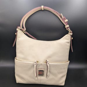 May include: A cream-colored leather hobo bag featuring a single adjustable shoulder strap with pink edges, a top zipper closure, and two front exterior pockets with zipper pulls. A rectangular metal logo plate is centered on the front exterior.