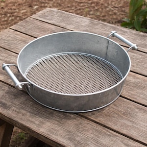 May include: A round, galvanized metal sifter with a fine mesh screen. The sifter has two metal handles and is sitting on a wooden surface. This gardening tool is used for sifting soil or compost.