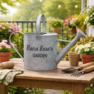 May include: A galvanized metal watering can with a handle and spout, inscribed with "Nana Rose's GARDEN." The can sits on a wooden table with gardening tools, gloves, and a ball of twine. Potted flowers in terracotta pots are in the background.