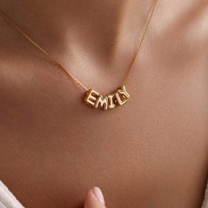 Könnte beinhalten: Eine goldfarbene Halskette mit dem Namen "EMILY", der in einzelnen Buchstabenperlen geschrieben ist. Die Halskette wird um den Hals getragen, wobei die Buchstaben in einer leichten Kurve hängen. Der Hintergrund ist unscharf.