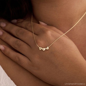 Puede incluir: Un delicado collar de oro con tres colgantes en forma de corazón. Cada corazón tiene una inicial grabada. El collar se muestra sobre la piel de una persona, resaltando el diseño. La cadena es fina y elegante, creando una joya personalizada.