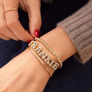 Puede incluir: Una colección de pulseras y un anillo dorados. Las pulseras incluyen una pulsera de cuentas, una pulsera fina con pequeñas piedras y una pulsera con la palabra "FRANKIE". El anillo es una fina banda de oro.