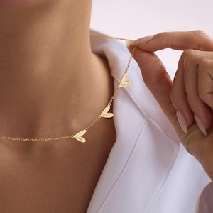 Puede incluir: Un collar dorado con tres colgantes en forma de corazón. Cada corazón tiene un nombre o fecha grabada. El collar lo sostiene una persona con una camisa blanca. Es una delicada pieza de joyería.