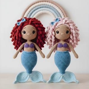 Op de afbeelding: Twee handgemaakte gehaakte zeemeerminnenpoppen. De ene pop heeft rood haar en de andere roze haar. Beide poppen hebben blauwe staarten, paarse tops en blauwe strikken. De poppen zijn ongeveer 30 cm hoog.