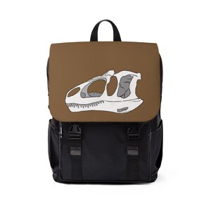Op de afbeelding: Een zwarte rugzak met een bruine klep voorzien van een witte en grijze illustratie van een dinosaurus-schedel. De tas heeft een handvat aan de bovenkant, twee zwarte verticale banden met kunststof gespen en zijvakken voor extra opbergruimte.