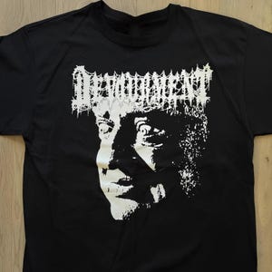 Könnte beinhalten: Ein schwarzes Kurzarm-T-Shirt mit einem weißen Grafikdruck auf der Brust. Das Design zeigt den Schriftzug "DEVOURMENT" in einer stilisierten, gezackten Schrift über einer kontrastreichen, schwarz-weißen Illustration eines Gesichts.