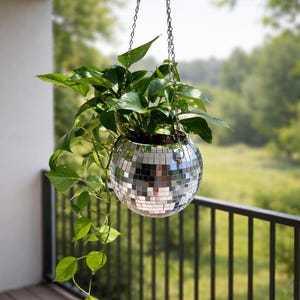 May include: A hanging disco ball planter filled with green leafy plants. The spherical planter is made of mirrored tiles and suspended by a silver chain. The background shows a wooden deck, black railing, and a blurred green landscape.