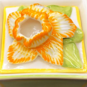 Puede incluir: Caja de baratijas cuadrada de cerámica con tapa con un diseño de narciso en 3D. La flor tiene pétalos naranjas y blancos con un centro amarillo, sobre un fondo blanco. La caja es de color verde claro con ribetes amarillos y blancos.