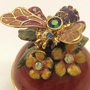 Op de afbeelding: Een decoratieve, rood geëmailleerde sieradendoos met een goudkleurige rand. De doos is bekroond met een gedetailleerde, goudkleurige bij en bloemversieringen. De bij heeft paarse en roze vleugels en is versierd met kleurrijke kristallen.