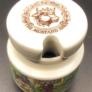 Pode incluir: Um pote de mostarda de cerâmica branca com tampa correspondente, apresentando um logótipo de cabeça de touro castanho e o texto THE MUSTARD SHOP. A base cilíndrica é decorada com uma ilustração gráfica colorida de figuras numa loja com fundo verde.