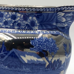 May include: A vintage blue and silver pitcher with a landscape design. The pitcher features a silver handle, rim, and base, with a blue body decorated with floral and scenic patterns. The design includes a house, trees, and flowers.