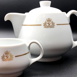 Könnte beinhalten: Weißes Porzellan-Teeservice mit Teekanne und Sahnekännchen. Beide Teile haben ein goldenes Emblem und ein goldenes Band. Die Teekanne hat einen Deckel und einen gebogenen Griff. Das Sahnekännchen hat einen Ausguss und einen kleinen Griff.