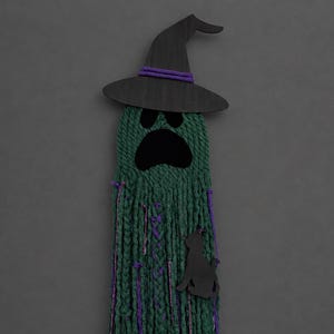 May include: A wall hanging decoration featuring a ghost figure made of green yarn strands. The ghost wears a black wooden witch hat with a purple yarn band. It has black felt eyes and a mouth, and a black cat silhouette attached to the green yarn.