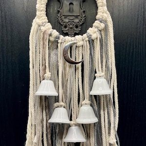 Op de afbeelding: Een wandhanger met een cirkelvormig macramé frame van dik gebroken wit katoenen koord, versierd met een zilveren maansikkelbedel, vijf witte keramische bellen en hangende strengen van gebroken wit touw en glinsterend zilver lint.