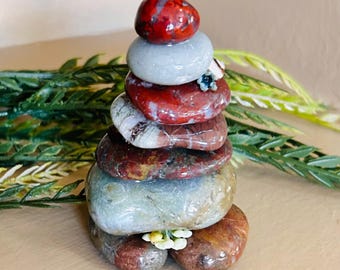 May include: A vertical stack of smooth, polished stones in varying colors including deep red, light gray, and mottled brown. Small decorative floral accents in blue and white are tucked between the layers of the stones, creating a balanced sculpture.