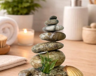 May include: A vertical stack of six smooth, rounded, earth-toned river stones arranged in a pyramid shape. The base is a flat, speckled stone adorned with small seashells, a tiny spiral shell, and a small green plant sprig at the front.