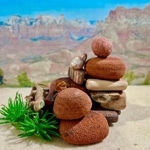 May include: A stacked arrangement of various stones and pebbles in shades of brown and tan. A small, tan lizard figurine rests near the base, with green plant-like accents. The background features a landscape of mountains and a blue sky.