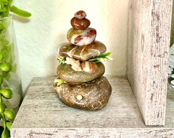 May include: A stack of five smooth, rounded river stones in shades of brown, tan, and white, arranged vertically. Small decorative white seashells and tiny green artificial leaves are tucked between the middle stones, creating a layered appearance.