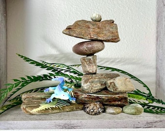 May include: A stack of smooth, earth-toned river stones arranged vertically, topped with a small, round, light-colored pebble. A small, bright blue plastic lizard figurine is positioned on a flat stone at the base, surrounded by green fern fronds.