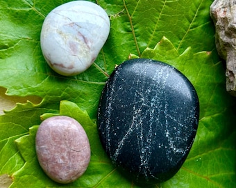 May include: Three smooth, polished stones in shades of white, pink, and black rest on a vibrant green leaf. The stones vary in size and shape, with the largest being a dark, speckled black. The leaf's texture and the stones' natural colors create a serene composition.