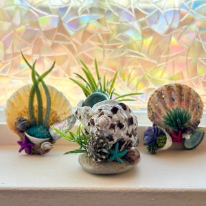 May include: A set of three ceramic mugs with a speckled, cream-colored glaze. Each mug features a different hand-painted design: a blue and green leaf, a yellow flower, and a red berry. The mugs have a simple, rounded shape.