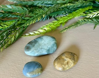 May include: Three polished stones in shades of blue, green, and tan. The stones have smooth, rounded shapes and varying patterns. Green leafy sprigs are in the background, creating a natural aesthetic.
