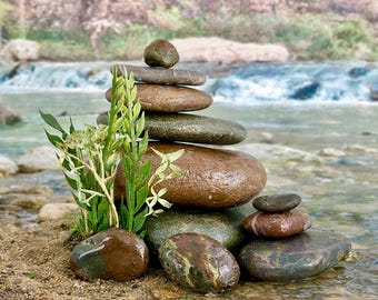 May include: A balanced stack of smooth, rounded river stones in varying shades of brown and gray, with a small plant to the left. The stones are arranged in a pyramid shape, with a natural water background.