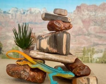 May include: A stack of smooth, multi-colored stones arranged in a cairn formation. A small, bright blue and orange plastic lizard figurine is positioned at the base of the stone stack, next to a small, spiky green artificial plant element.