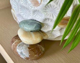 May include: A stack of four polished stones in shades of brown, tan, and blue-green. The stones are arranged in a balanced tower, with a white ceramic vase with a floral pattern in the background. Green leaves are in the upper right corner.