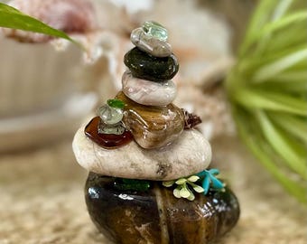May include: A vertical stack of smooth, polished stones in various shades of brown, gray, and white. The arrangement includes small, translucent green and amber glass pieces, along with tiny artificial white and blue floral accents.