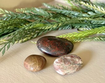 May include: Three polished stones in shades of brown, red, and pink are displayed. The stones have smooth, rounded shapes and varying patterns. Green and yellow leafy sprigs are in the background. The stones are likely used for decorative or healing purposes.