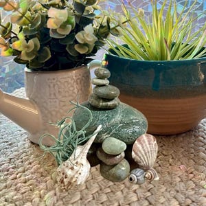 May include: A zen-inspired still life featuring a stack of smooth, gray stones, seashells, and small plants in pots. The scene is set on a woven mat, with a watering can and a window in the background.