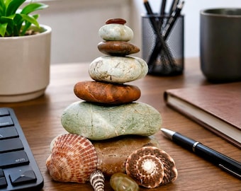 May include: A stack of six smooth, rounded river stones in varying shades of gray, green, and brown, arranged vertically. At the base of the stone tower are two spiral seashells, one small conical shell, and one additional small green pebble.