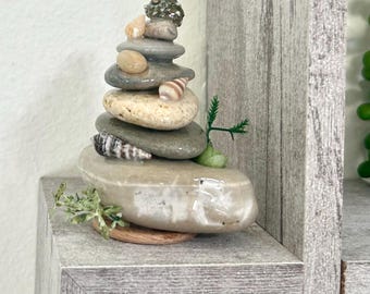 May include: A decorative stack of smooth, rounded river stones arranged vertically. The stones are interspersed with small, varied seashells in shades of tan, brown, and white. A tiny green artificial plant sprig is tucked into the side of the base.