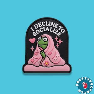 May include: A black patch with the text "I DECLINE TO SOCIALIZE" in white. The patch features a green frog wrapped in a pink blanket with pink hearts, stars, and a flower. The patch is a fun accessory.