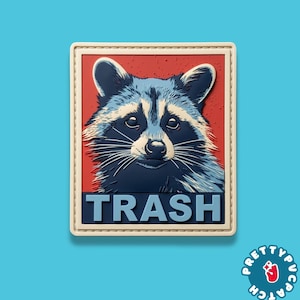 May include: A rectangular PVC patch featuring a stylized illustration of a raccoon face against a red background. The word TRASH is printed in large, light blue capital letters at the bottom. The patch has a cream-colored border with stitching.