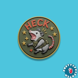 May include: A round patch featuring a cartoon opossum wearing a tactical vest and yelling. The word "HECK" is arched above the opossum in orange letters. The patch has a green background with gold stars and a gold border.