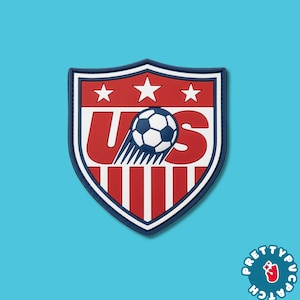 May include: A shield-shaped patch featuring the letters US in red with a blue and white soccer ball replacing the letter S. The design includes three white stars at the top and vertical red and white stripes at the bottom, all outlined in dark blue.