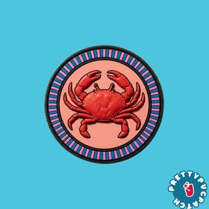 May include: A circular patch featuring a raised red crab centered on a light peach background. The border consists of alternating vertical blue and pink stripes enclosed by a thick black ring. The design is flat and symmetrical in its layout.
