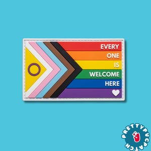 May include: A rectangular patch featuring the Progress Pride flag design on the left side and horizontal stripes of red, orange, yellow, green, blue, and purple on the right. The text EVERY ONE IS WELCOME HERE is printed in white with a small white heart.