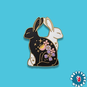 May include: A black and white enamel pin featuring two rabbits facing each other. The black rabbit has gold stars and floral details in pink and purple. The pin has a gold outline and is set against a blue background. The pin has the text "PRETTYPUCPA" in a circle.