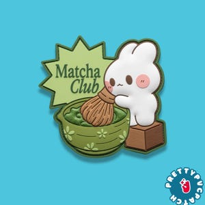 May include: A white bunny character with pink cheeks and a brown whisk stirring matcha in a green bowl. The image includes the text "Matcha Club" on a green starburst shape. The background is a solid light blue.