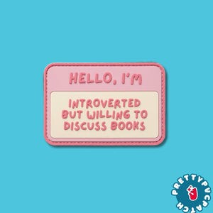 May include: A rectangular name tag patch with a pink border and a cream-colored center. The text HELLO, I'M is printed in pink at the top, followed by the text INTROVERTED BUT WILLING TO DISCUSS BOOKS in pink lettering in the center of the patch.